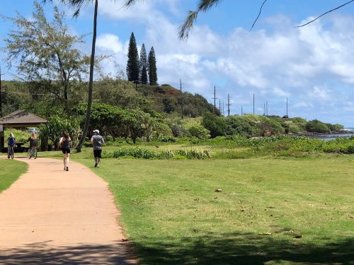 A scenic pathway with people walking, surrounded by greenery and trees, under a blue sky with fluffy clouds.