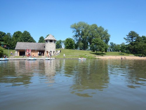 A serene lake scene with a sandy beach, green trees, and a stone building under a clear blue sky.
