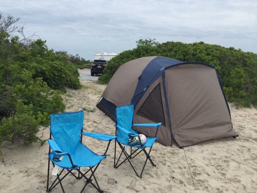 A beach scene featuring a tent and two blue folding chairs on sandy ground, surrounded by greenery.