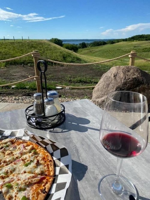 A scenic view of a lake and hills with a pizza and a glass of red wine on a table in the foreground.