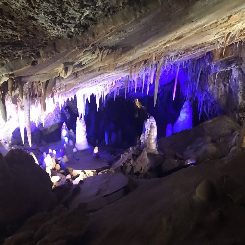 A dimly lit cave with stalactites and stalagmites, illuminated by purple and blue lights.