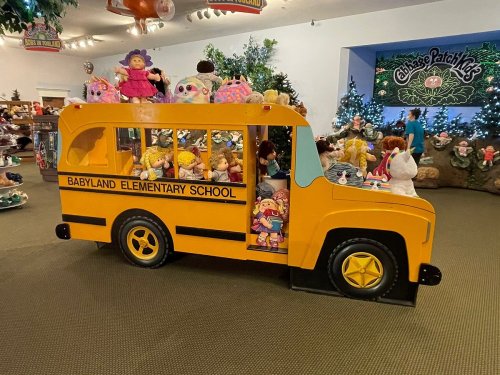 A colorful toy school bus filled with dolls, set in a festive display with holiday decorations in the background.
