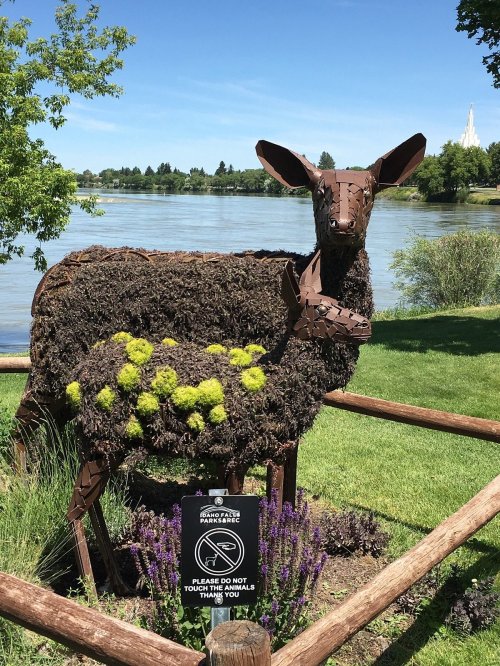 A large sculpture of a deer made from natural materials, standing by a river with greenery and flowers around it.