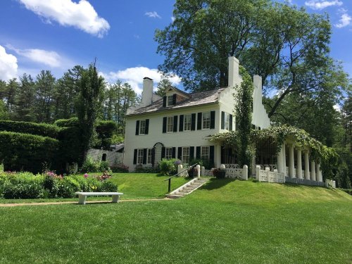A charming white house with green shutters, surrounded by lush gardens and trees under a bright blue sky.