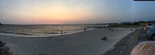 A panoramic view of a beach at sunset, with people walking along the shore and gentle waves lapping at the sand.