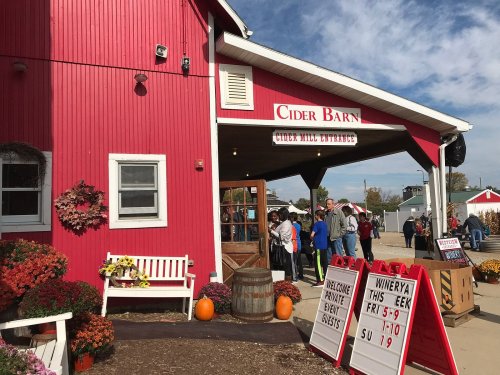 A red barn with a sign for "Cider Mill Entrance," surrounded by people and autumn decorations like pumpkins and flowers.