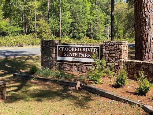 Sign for Crooked River State Park in Georgia, surrounded by trees and greenery, with a road visible in the background.