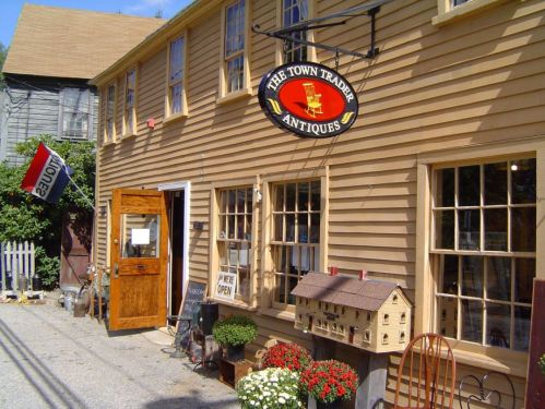 A quaint antique shop with a wooden facade, open sign, and flower pots outside, set in a charming street.