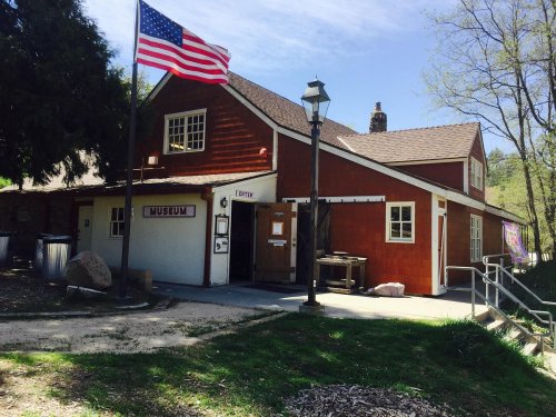 A red wooden building with a sign reading "Museum," an American flag, and a lamp post in a grassy area.