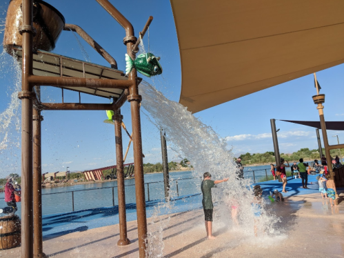 Children play under a large water feature, with water cascading down from a bucket in a sunny outdoor setting.