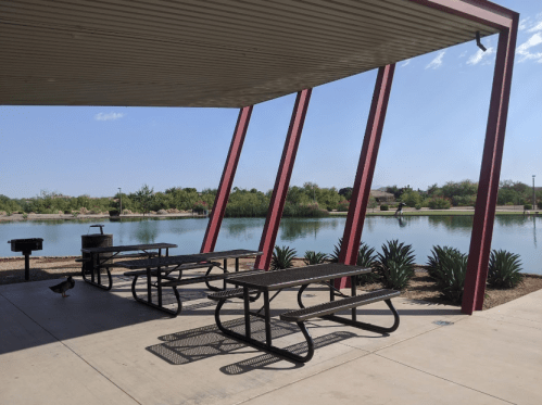 A shaded picnic area with metal tables overlooks a calm pond surrounded by greenery and blue skies.
