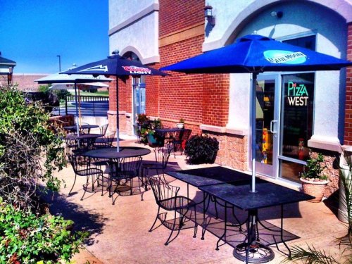Outdoor seating area with black tables and umbrellas outside a restaurant, surrounded by plants and a clear blue sky.