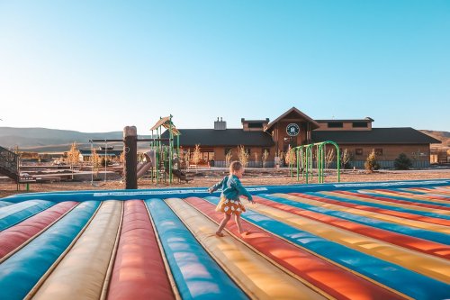 A child runs on a colorful inflatable surface with a playground and building in the background under a clear blue sky.