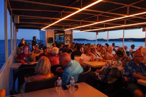 A lively crowd enjoys an evening on a boat, with music and drinks, as the sun sets over the water.