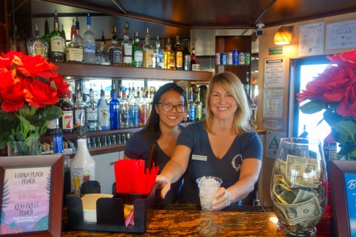 Two women smile behind a bar filled with bottles, surrounded by red flowers and a money jar. One holds a drink.