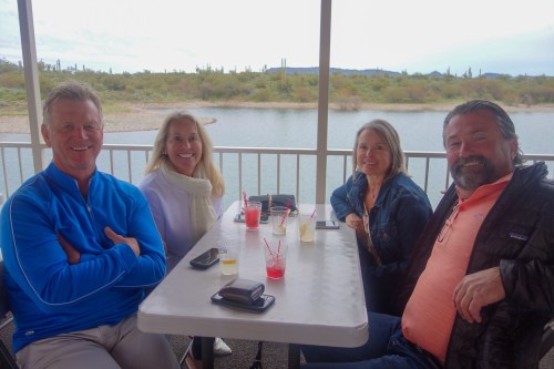 A group of four friends sitting at a table by a lake, enjoying drinks and smiling at the camera.