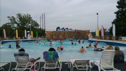 A busy wave pool at Point Mallard, with people swimming and lounging on chairs under a cloudy sky.
