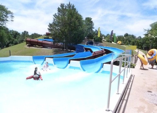 A water park scene featuring blue water slides, a pool, and people enjoying the water on a sunny day.