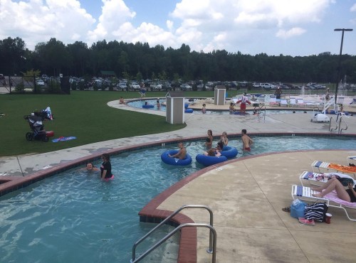 A pool area with people swimming, lounging, and floating on tubes, surrounded by green grass and trees under a cloudy sky.