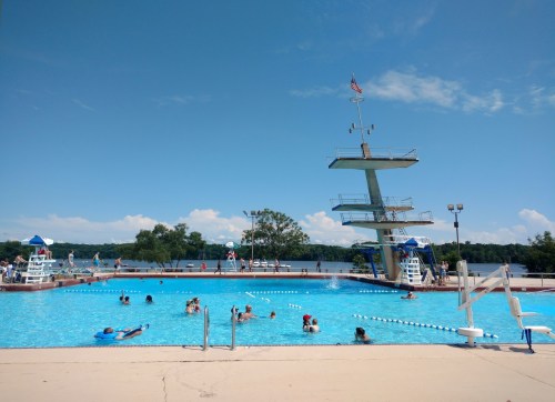 A sunny outdoor pool with a diving board, people swimming, and trees in the background under a clear blue sky.