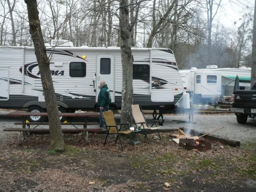 A campsite with a camper, a man standing near a fire pit, and picnic tables surrounded by trees.