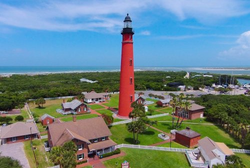 Aerial view of a tall red lighthouse surrounded by green trees and buildings near the coast under a blue sky.