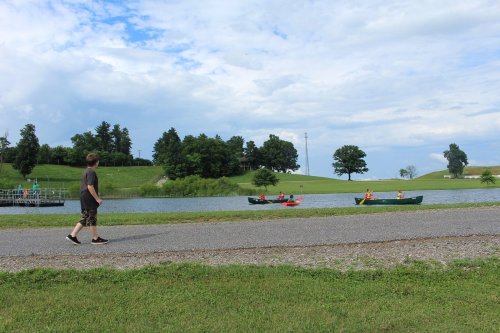 A person walks along a path by a lake where others are kayaking, surrounded by green hills and trees under a cloudy sky.