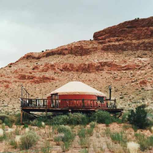 A cozy yurt on a wooden deck, set against a backdrop of red rock formations and sparse vegetation.