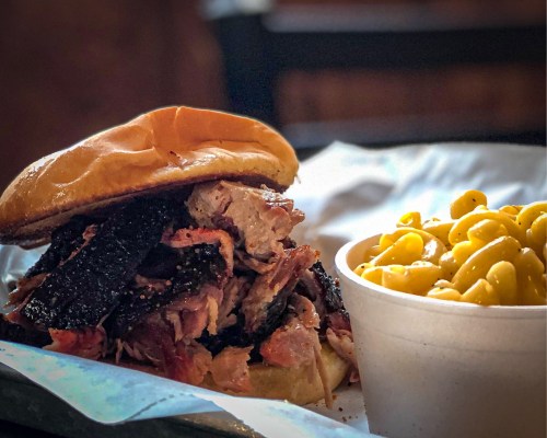 A close-up of a pulled pork sandwich with a soft bun, served alongside a cup of creamy macaroni and cheese.