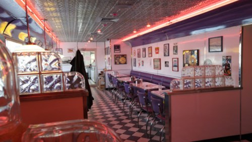 A retro diner interior with checkered floors, neon lights, and vintage decor on the walls.