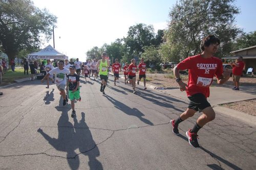 A group of children and teens running in a race on a sunny day, with a tent in the background.