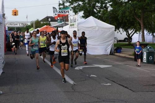 A group of runners in athletic gear races down a street lined with tents, with a finish line banner in the background.