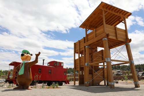 A large cartoon bear statue waves next to a wooden lookout tower and a red train car under a cloudy sky.