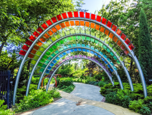 A colorful archway made of transparent panels in rainbow colors, surrounded by lush greenery and a winding pathway.