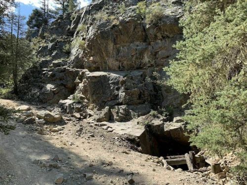 A rocky hillside with trees, featuring an old mine entrance partially obscured by dirt and rocks.