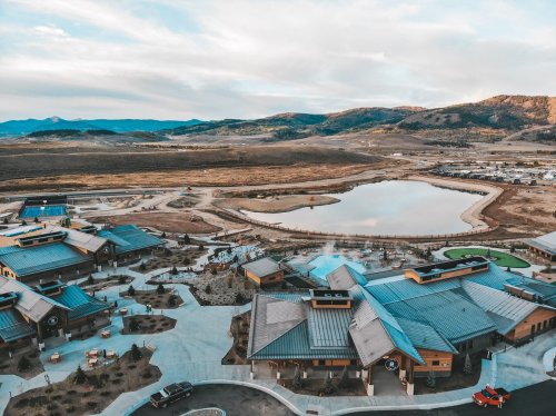 Aerial view of a resort with modern buildings, a pond, and surrounding mountains under a cloudy sky.