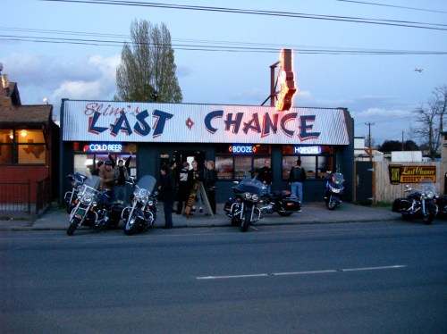 A group of people stands outside a bar named "Last Chance," with motorcycles parked nearby and a neon sign glowing.