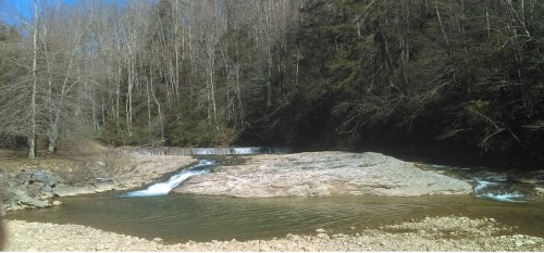 A serene river scene with flowing water over rocks, surrounded by trees and a clear blue sky.