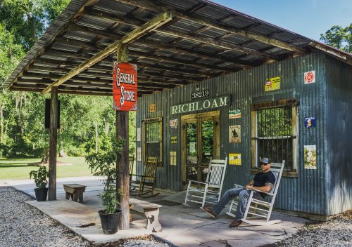 A rustic general store with a metal roof, rocking chairs, and vintage signs, surrounded by greenery. A man sits outside.
