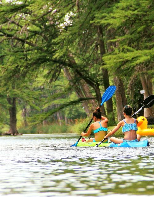 Two girls in bikinis kayak on a serene river surrounded by lush green trees.