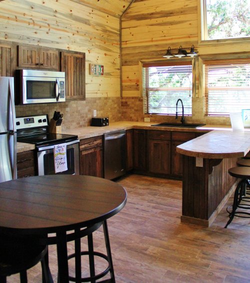 Cozy kitchen with wooden walls, modern appliances, and a small dining table. Bright natural light from the windows.