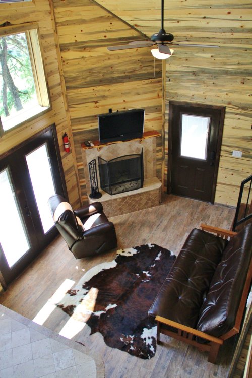Cozy living room with wooden walls, a fireplace, a leather couch, and a cowhide rug, featuring large windows and a door.
