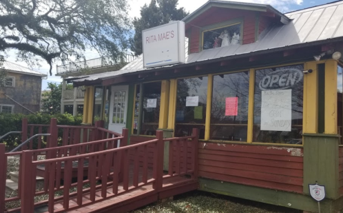 A colorful restaurant with a sign reading "Rita Mae's," featuring a ramp and large windows, surrounded by greenery.