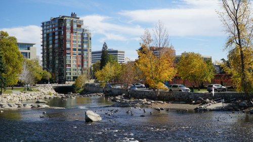 A river flows past a city park with autumn trees and a modern building in the background under a clear blue sky.