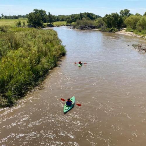 Two kayakers paddle down a calm river surrounded by lush greenery and a clear blue sky.