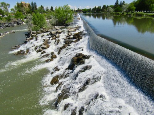 A serene river scene with a waterfall cascading over rocks, surrounded by lush greenery and clear blue skies.