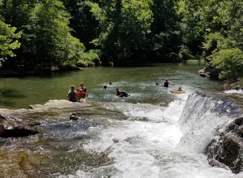 People swimming and playing in a serene river surrounded by lush greenery and a small waterfall.