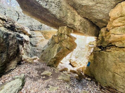 A rocky cave with large boulders and a natural stone arch, surrounded by earthy tones and scattered leaves.