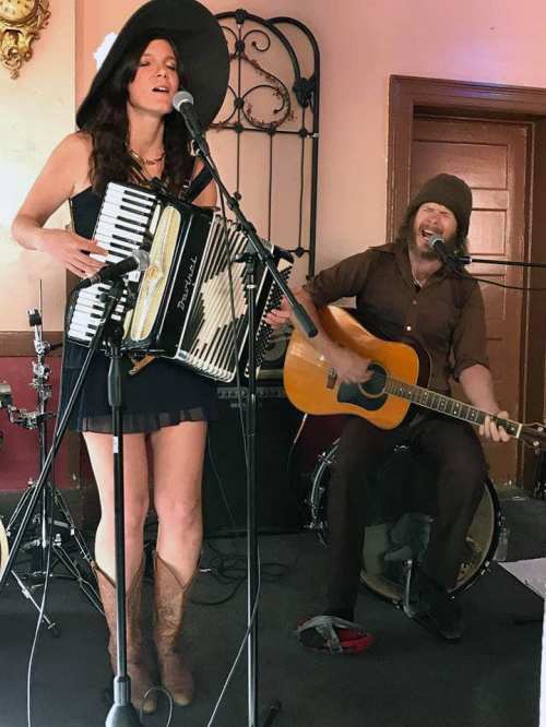 A woman in a black dress and hat plays the accordion while a man in a brown hat strums an acoustic guitar on stage.