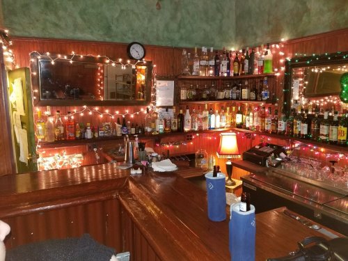 A cozy bar with a wooden counter, shelves lined with various bottles, and festive lights creating a warm atmosphere.